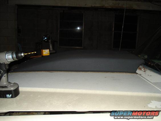 Drill bit poised above a dark-colored vehicle roof section. A yellow box sits behind the drill