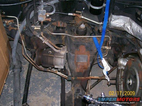 Engine bay components including AC compressor, hoses, and suspension parts, with a blue strap attached