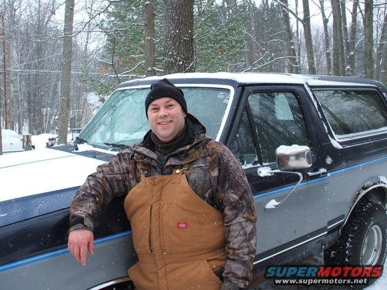 Man in camouflage jacket and brown overalls leans on a 1990 Ford Bronco in the snow