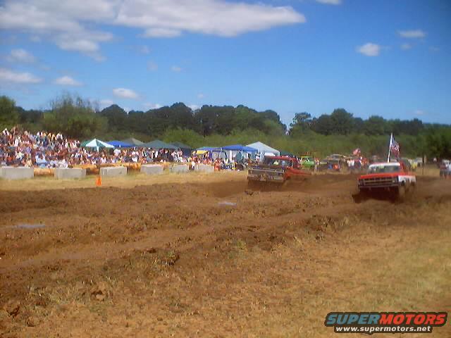 Mud bog race with multiple trucks competing, spectators in background