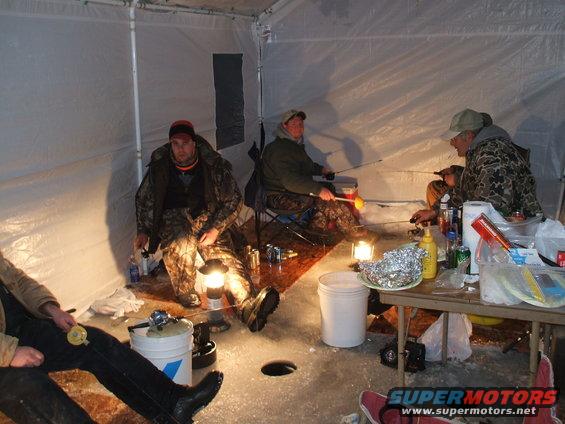 Four people ice fishing inside a tent, with fishing rods, lanterns, and food on a table