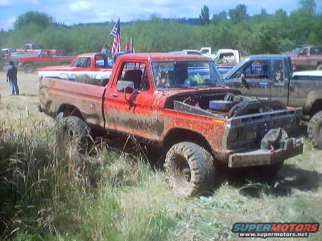 Mud-covered red Ford pickup truck with hood open, parked in grassy field