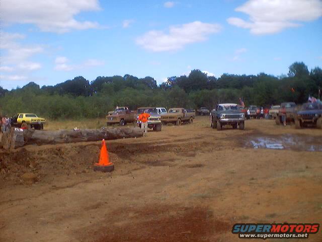 Mud drag racing event with several trucks on a dirt track under a cloudy sky. A large log is in the foreground