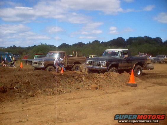 Two trucks stuck in mud at an event. One is a dark colored Ford F-100, the other is a brown Ford F-100