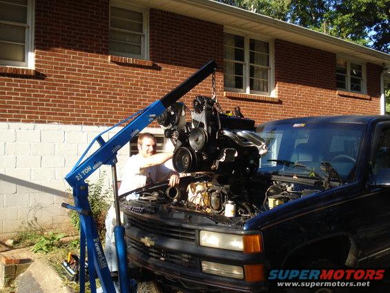 Guy using engine hoist to lift engine out of dark blue pickup truck in front of brick house