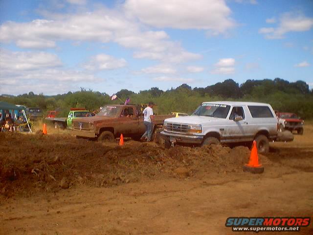 Muddy trucks and SUVs stuck in the mud at an event. A white SUV is in the foreground, with a rusty pickup truck behind it