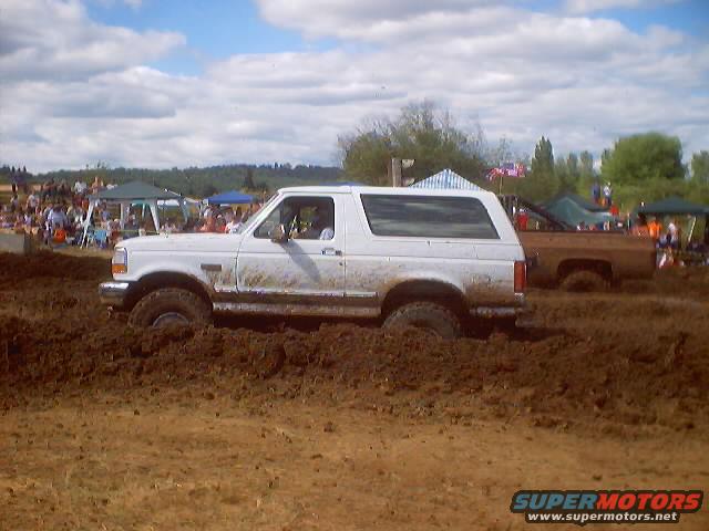 White Ford Bronco stuck in mud, side view, with a brown pickup truck behind it