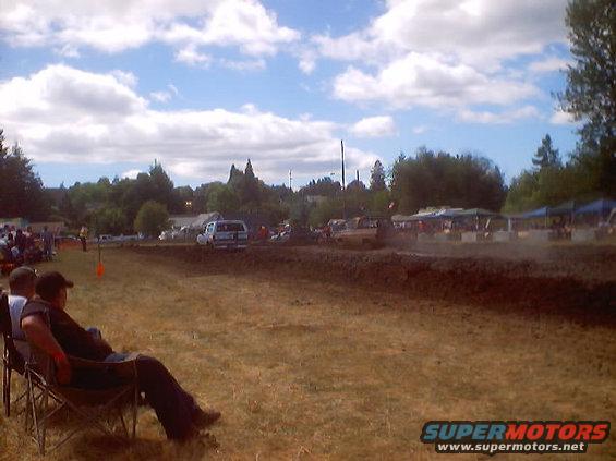 Mud drag racing event with spectators in chairs and vehicles on a dirt track under a cloudy sky