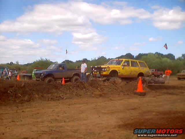 Muddy field with a blue pickup truck and a yellow station wagon. Orange cones are on the ground