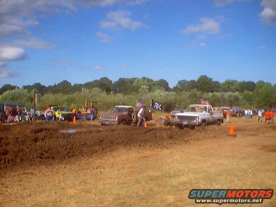 Mud bog event with several trucks stuck in mud, spectators watching under a blue sky