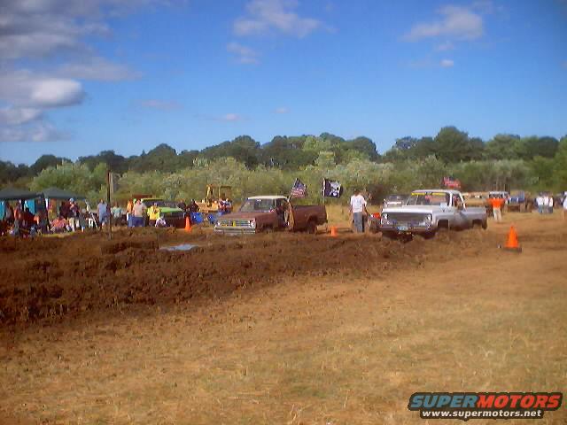 Mud bogging event with several trucks in the mud, spectators under tents, and trees in the background