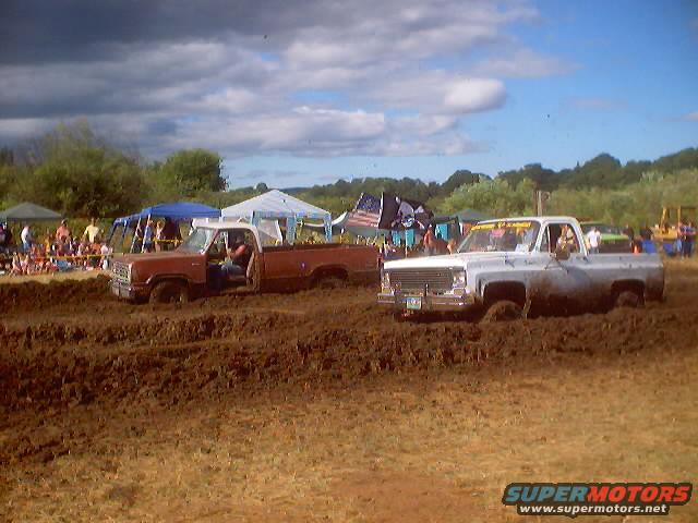 Two mud-splattered pickup trucks stuck in a mud bog, with spectators and tents in the background