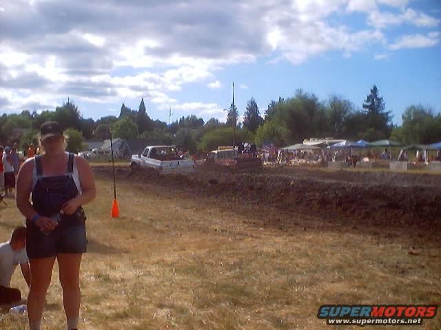 Mud drag race event with a white truck stuck in the mud, spectators in the background, and a woman in overalls in the foreground