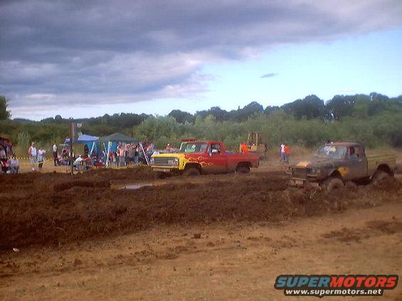 Two trucks stuck in mud at an outdoor event with spectators and tents in the background