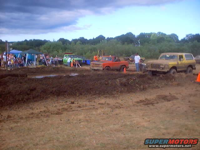 Mud bog event with multiple trucks stuck in mud, spectators under tents, and trees in background. Yellow Ford Bronco and orange Ford pickup visible