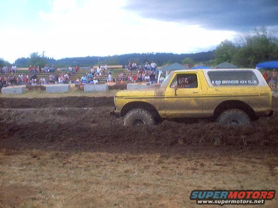 Yellow Ford Bronco stuck in mud pit at an event, with spectators in the background