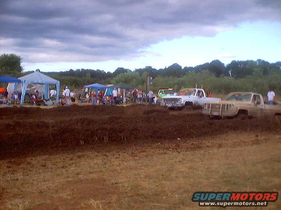 Two trucks stuck in mud at a drag race, one white, one tan. A crowd watches under tents