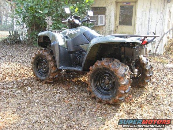 Olive green 2007 Suzuki King Quad 700 ATV with large, muddy tires parked on a dirt and leaf-covered surface