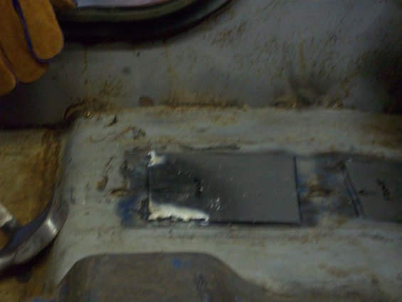 Close-up of a metal patch being welded onto a rusty vehicle floor. A gloved hand is visible at the top