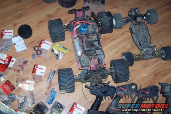 Two disassembled radio-controlled cars with numerous parts and controllers scattered on a wooden floor