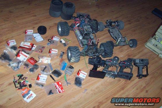 Two remote control cars, several controllers, and a pile of parts laid out on a wooden floor