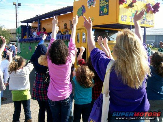 Yellow Mardi Gras float with people waving, text reads 'OF THE BAYOU'