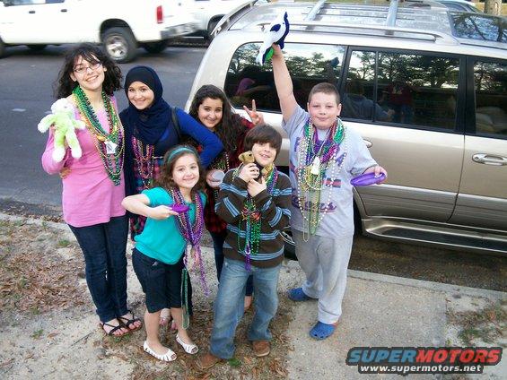 Group of kids in Mardi Gras beads with a white SUV and a gold SUV in the background