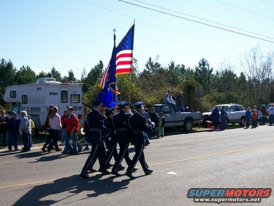 Color guard marches in a parade past spectators and an RV, pickup truck, and SUV