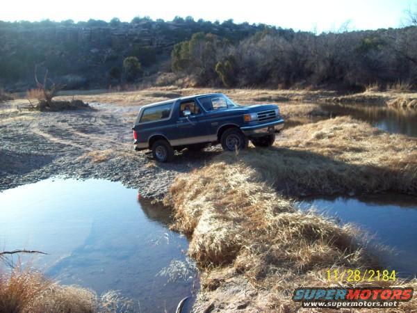Blue 1990 Ford Bronco driving on a dirt path next to a body of water