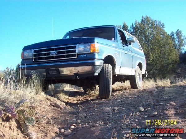 Blue 1990 Ford Bronco driving on a rocky dirt road with trees and blue sky