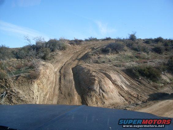 Off-road trail with tire tracks on a dry, scrub-covered hillside under a clear blue sky. Part of a dark vehicle hood is visible in the foreground