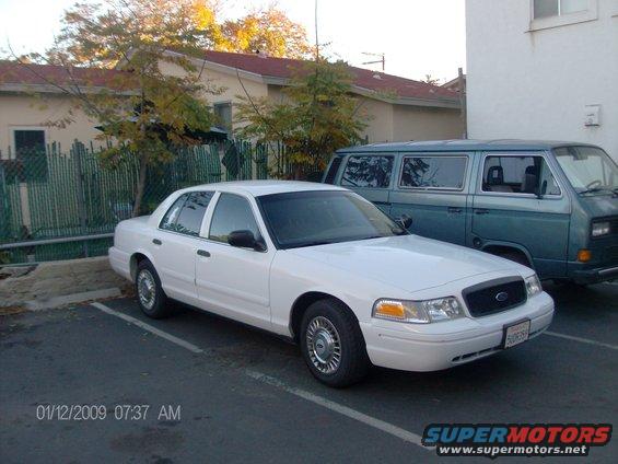 White sedan parked in a lot, blue van behind it. License plate visible