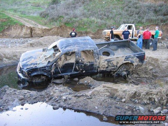 Black pickup truck submerged in mud with broken windshield and missing doors, another tan truck behind it