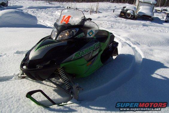 Green and black snowmobile with orange number 14 on windshield, parked in snow. Trucks in background