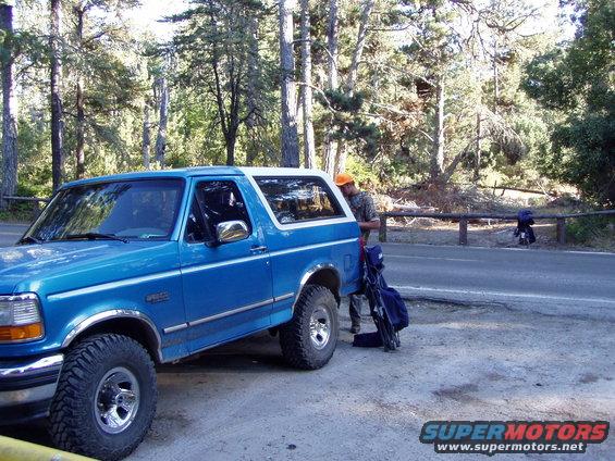 Blue 1995 Ford Bronco parked on a dirt shoulder next to a road, a person in an orange hat loads gear into the back