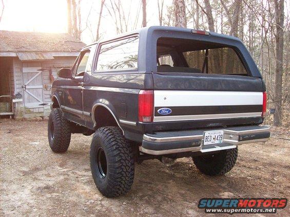 1992 Ford Bronco lifted with large off-road tires, parked on dirt near a wooden shed