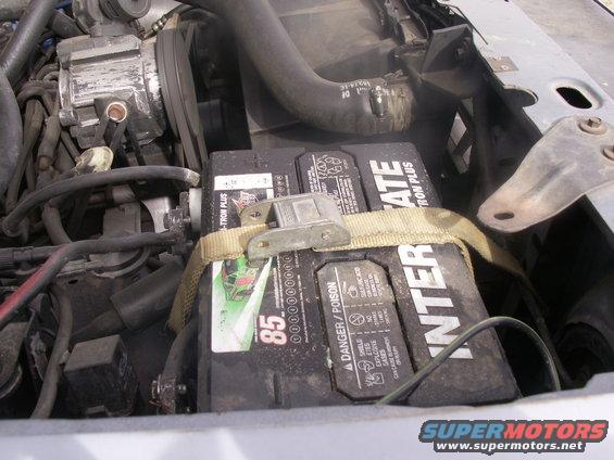 Engine bay showing a car battery secured with a strap, surrounded by hoses and engine components