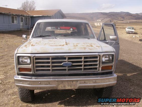 1986 Ford Bronco front view, door open. Red car visible in windshield reflection. White truck in background