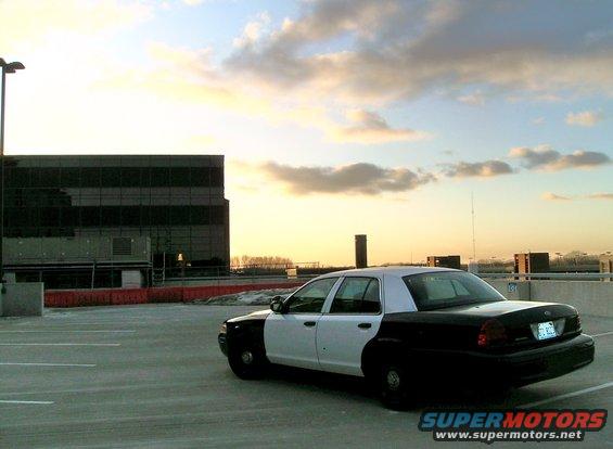 Two-tone police car, white front and black rear, parked in a lot with a modern building and cloudy sky