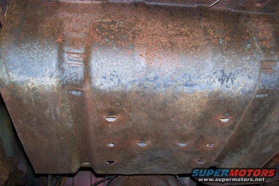Heavily rusted metal undercarriage part with visible indentations and holes. Some blueish discoloration present