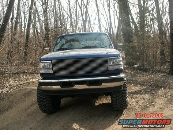 Blue Ford F-Series truck with aftermarket grille parked on a dirt road in woods