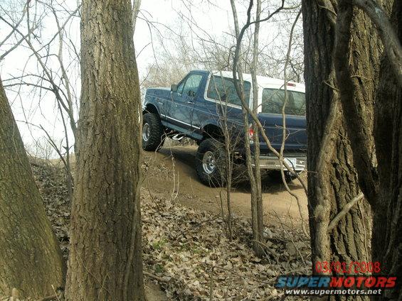 Blue Ford Bronco with a white top, lifted with large tires, driving on a dirt path