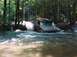 1993 Ford Bronco - An SUV with a red roof and silver body navigates a shallow stream in a wooded area.