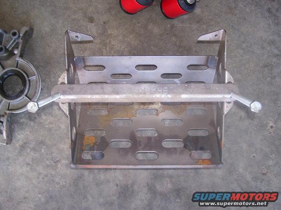 Metal battery tray with slotted holes and a crossbar, next to a silver engine component