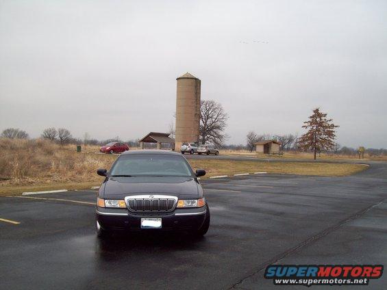 Dark sedan parked on wet asphalt, front view. Silo and buildings in background