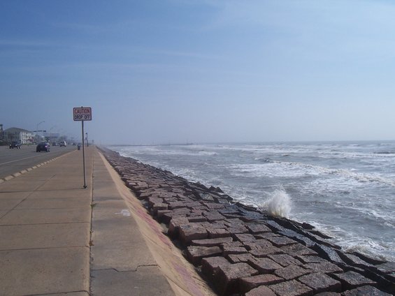 road next to a stone sea wall with waves crashing, caution drop off sign visible