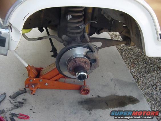 close-up of a vehicle's front wheel hub, brake rotor, and suspension components, with an orange floor jack supporting it