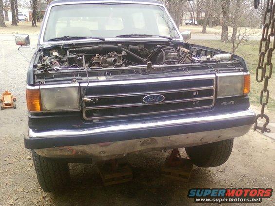 Ford Bronco with hood open, engine exposed. Front grille has Ford logo and 4x4 badge. Vehicle is on jack stands
