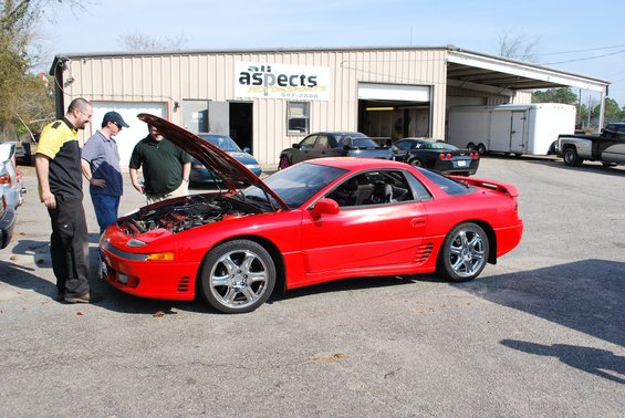 Red 1991 Mitsubishi 3000GT with hood open, three men looking at engine. Building in background