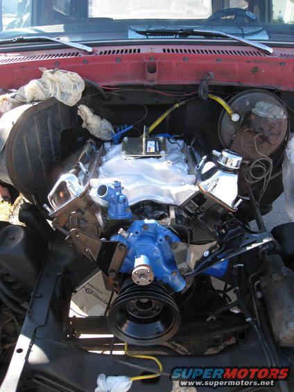Engine bay with a blue V8 engine, chrome components, and various hoses. Some white cloths are stuffed into openings